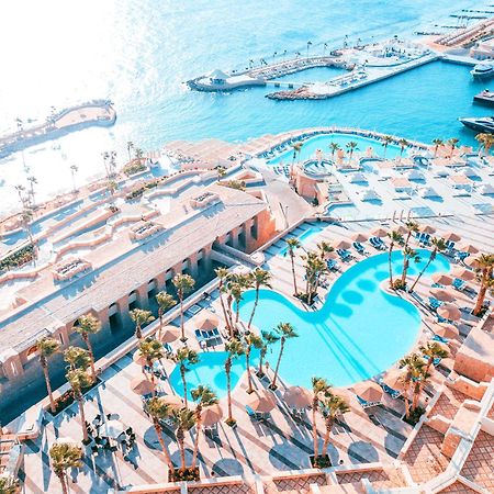 Pickalbatros Citadel Resort Sahl Hasheesh (Adults Only) Hurghada Exterior foto The photo shows a luxurious resort with a large, shaped swimming pool surrounded by palm trees and lounge chairs. The pool has a unique curve design, enhancing the overall aesthetic. In the background, there's a beautiful waterfront view with boats a