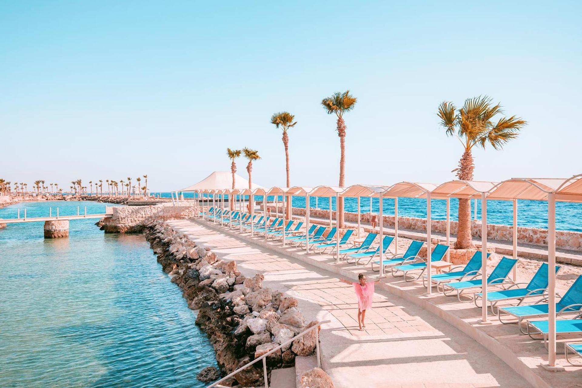 Pickalbatros Citadel Resort Sahl Hasheesh (Adults Only) Hurghada Exterior foto The photo depicts a serene seaside area characterized by a clear blue sky and calm waters. There are several palm trees lining a path beside the water, which leads to a row of beach chairs. The beach chairs are arranged under sunshades, creating a re