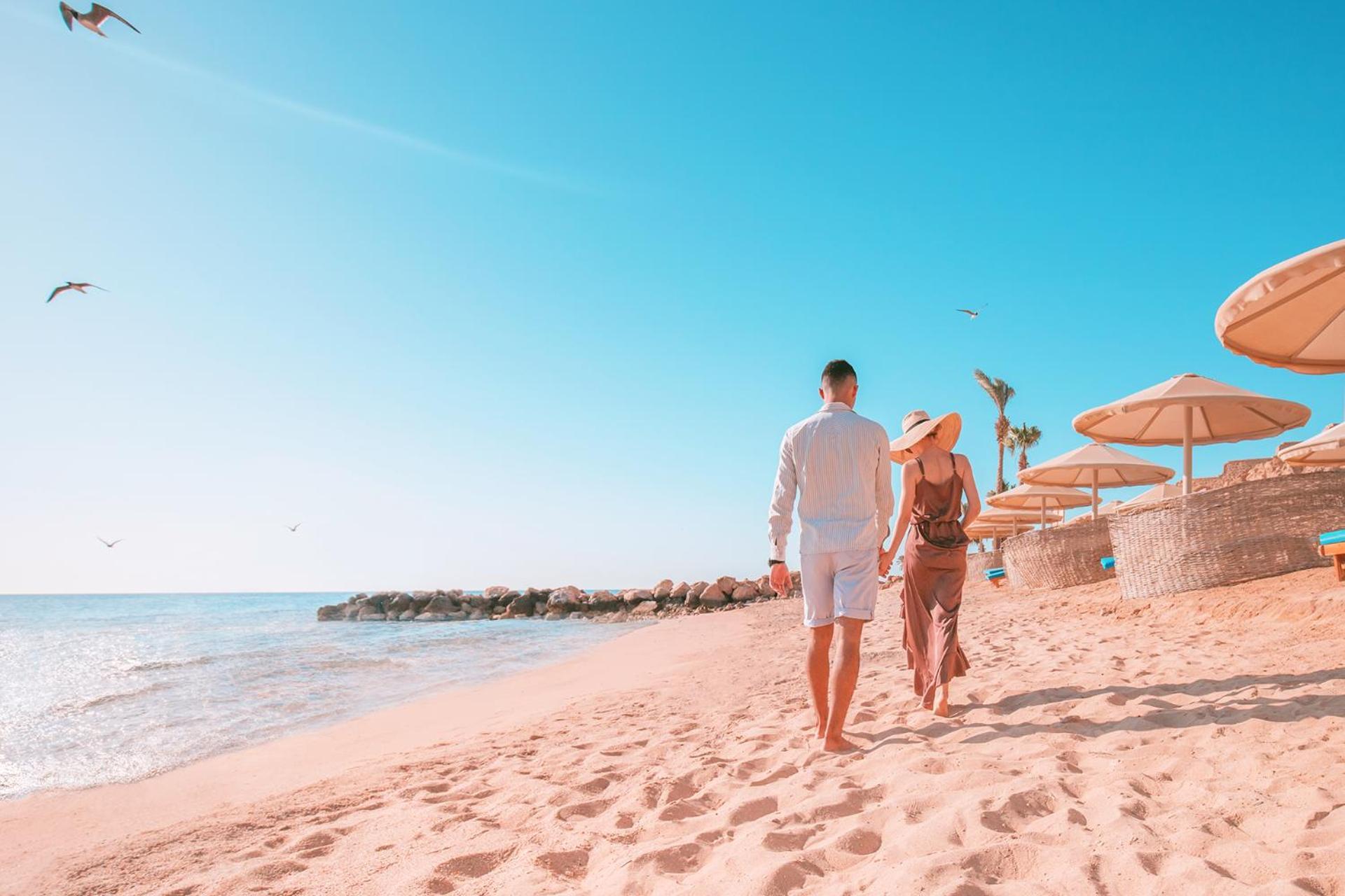 Pickalbatros Citadel Resort Sahl Hasheesh (Adults Only) Hurghada Exterior foto The photo shows a couple walking hand in hand along a sandy beach. They are facing the ocean, with the sun shining brightly. The woman is wearing a straw hat and has a relaxed outfit, while the man is dressed in a light-colored shirt and shorts. In t