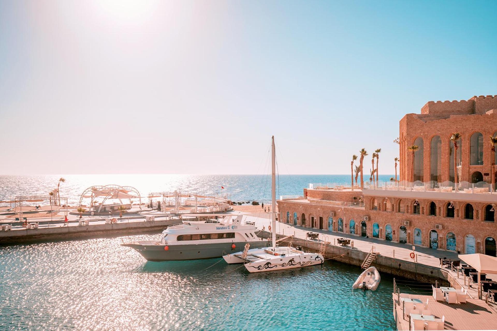 Pickalbatros Citadel Resort Sahl Hasheesh (Adults Only) Hurghada Exterior foto The photo depicts a picturesque harbor scene. In the foreground, several boats are docked in the calm, shimmering water under a bright blue sky. One of the vessels is a larger yacht, while smaller boats are also present. In the background, there are 