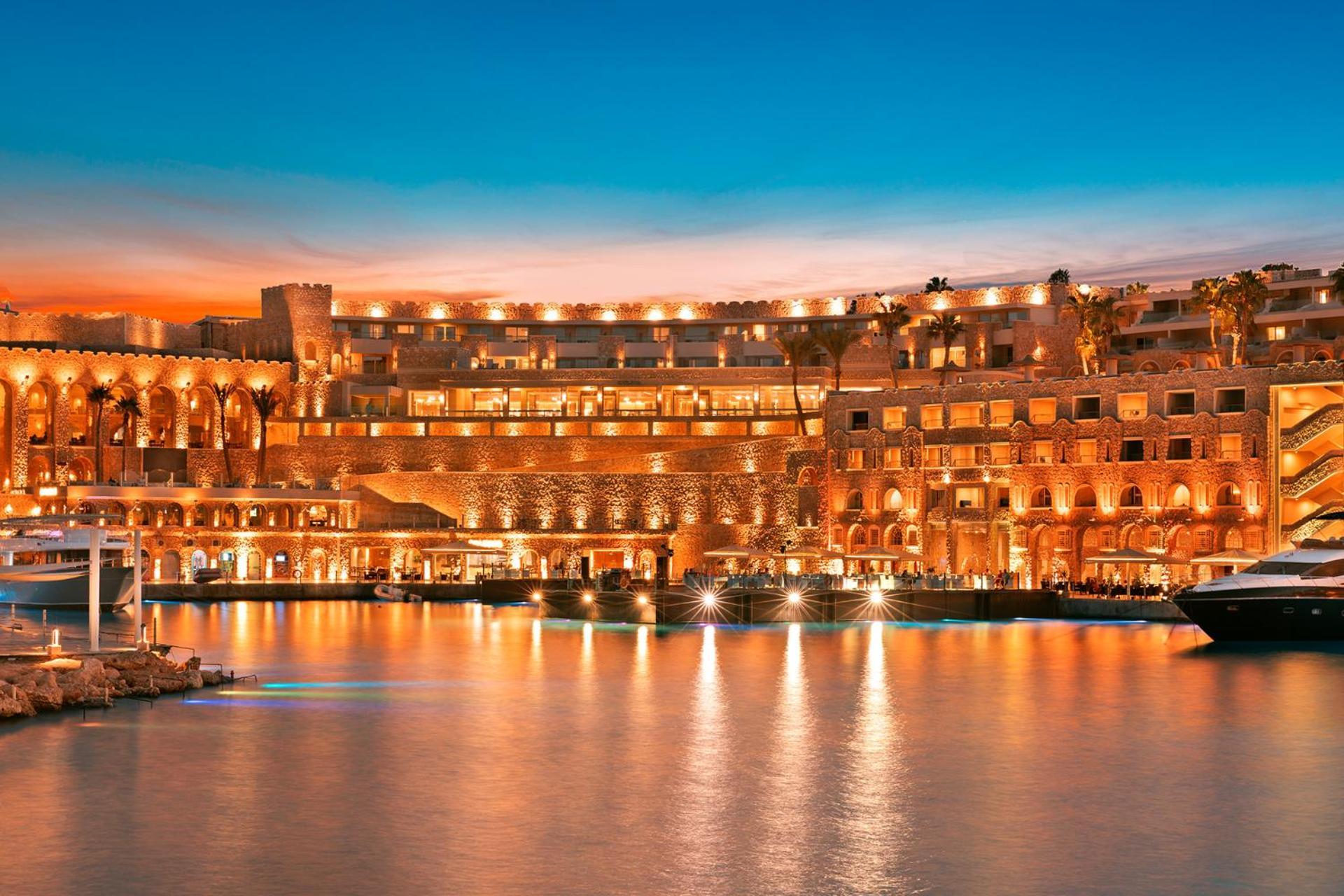 Pickalbatros Citadel Resort Sahl Hasheesh (Adults Only) Hurghada Exterior foto The photo depicts a luxurious waterfront resort or hotel illuminated during sunset. The building features a blend of modern and traditional architectural styles, with an array of balconies, terraces, and large windows that reflect the warm evening li