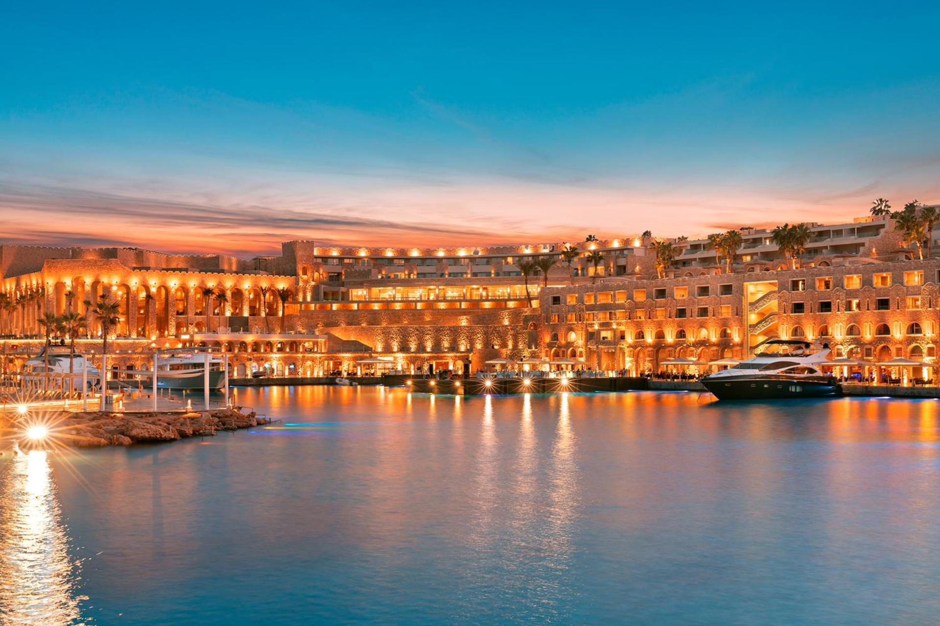 Pickalbatros Citadel Resort Sahl Hasheesh (Adults Only) Hurghada Exterior foto The photo depicts a beautiful waterfront scene during sunset. On the right, there is a luxurious building illuminated with warm lights, reflecting on the calm water. You can see boats moored in front of the structure, contributing to a tranquil ambia
