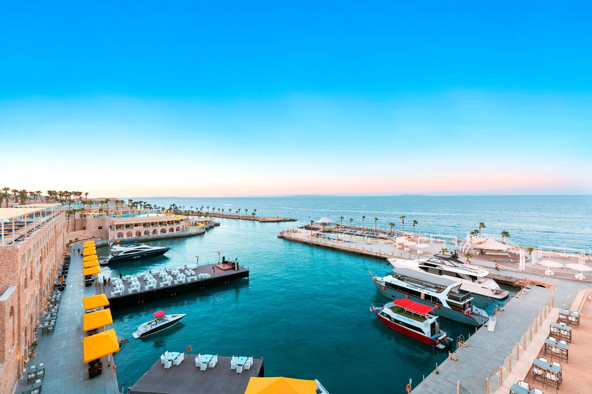 Pickalbatros Citadel Resort Sahl Hasheesh (Adults Only) Hurghada Exterior foto The photo depicts a serene harbor scene by the sea. There are various boats docked, including a few larger yachts and smaller vessels. The water is a vibrant blue, reflecting the clear sky above, which transitions from light blue to a warmer hue near