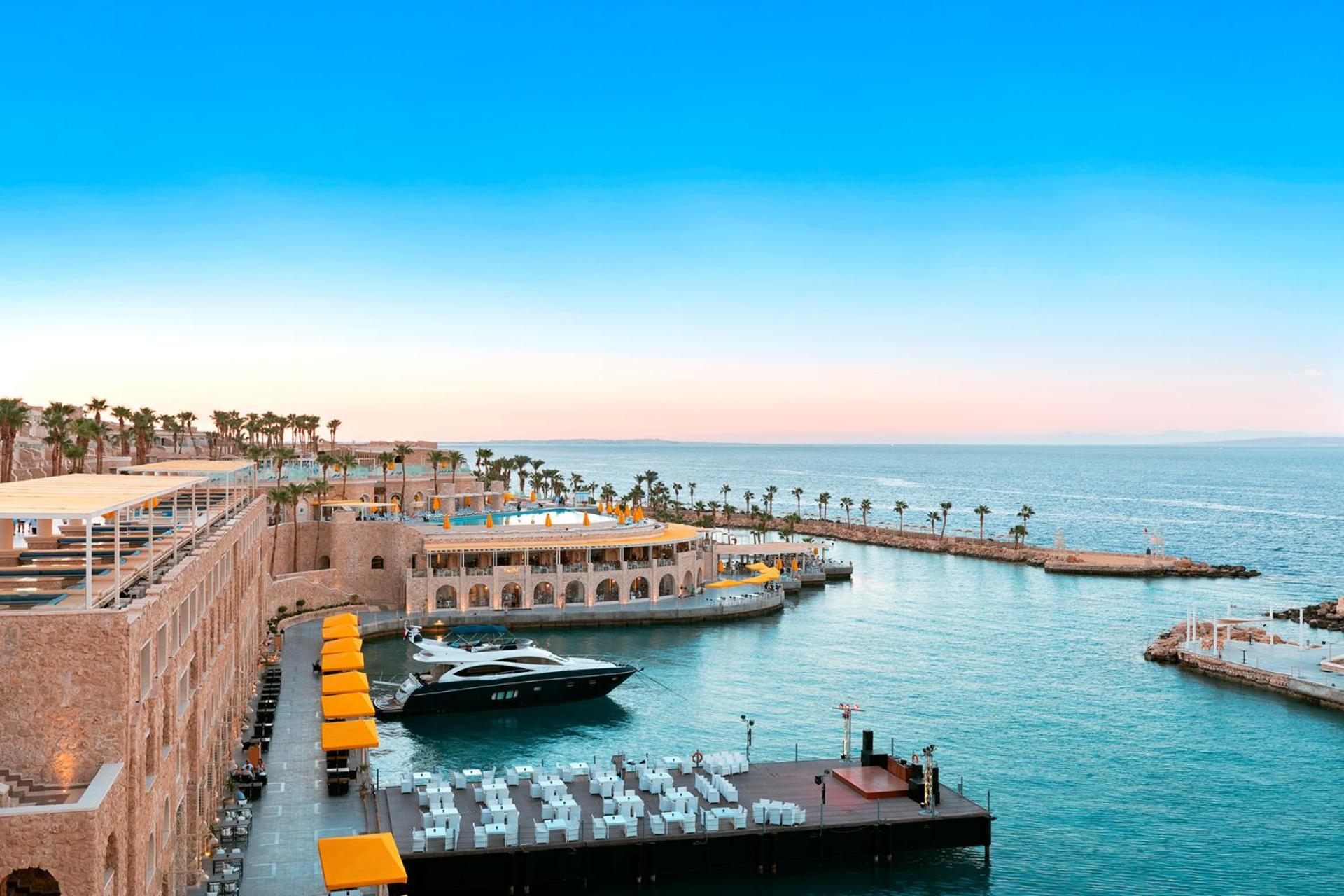 Pickalbatros Citadel Resort Sahl Hasheesh (Adults Only) Hurghada Exterior foto The photo depicts a picturesque waterfront scene. In the foreground, there is a marina with a sleek yacht docked alongside a floating platform lined with white tables and chairs. Beyond the marina, a large building features arched windows and outdoor