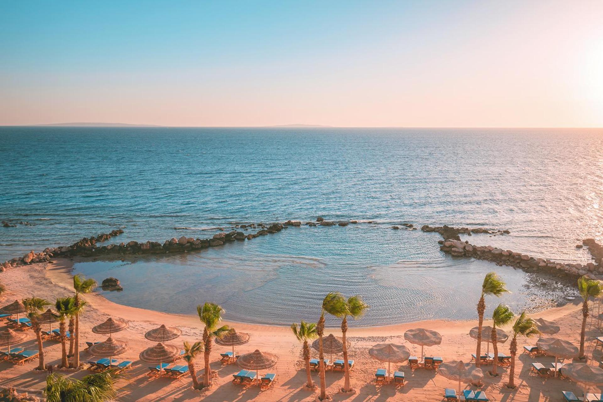 Pickalbatros Citadel Resort Sahl Hasheesh (Adults Only) Hurghada Exterior foto The photo depicts a serene beach scene. In the foreground, there are sandy areas with palm trees and thatched umbrellas, suggesting a tropical environment. Lounging chairs are positioned on the beach, inviting relaxation. In the background, the calm 