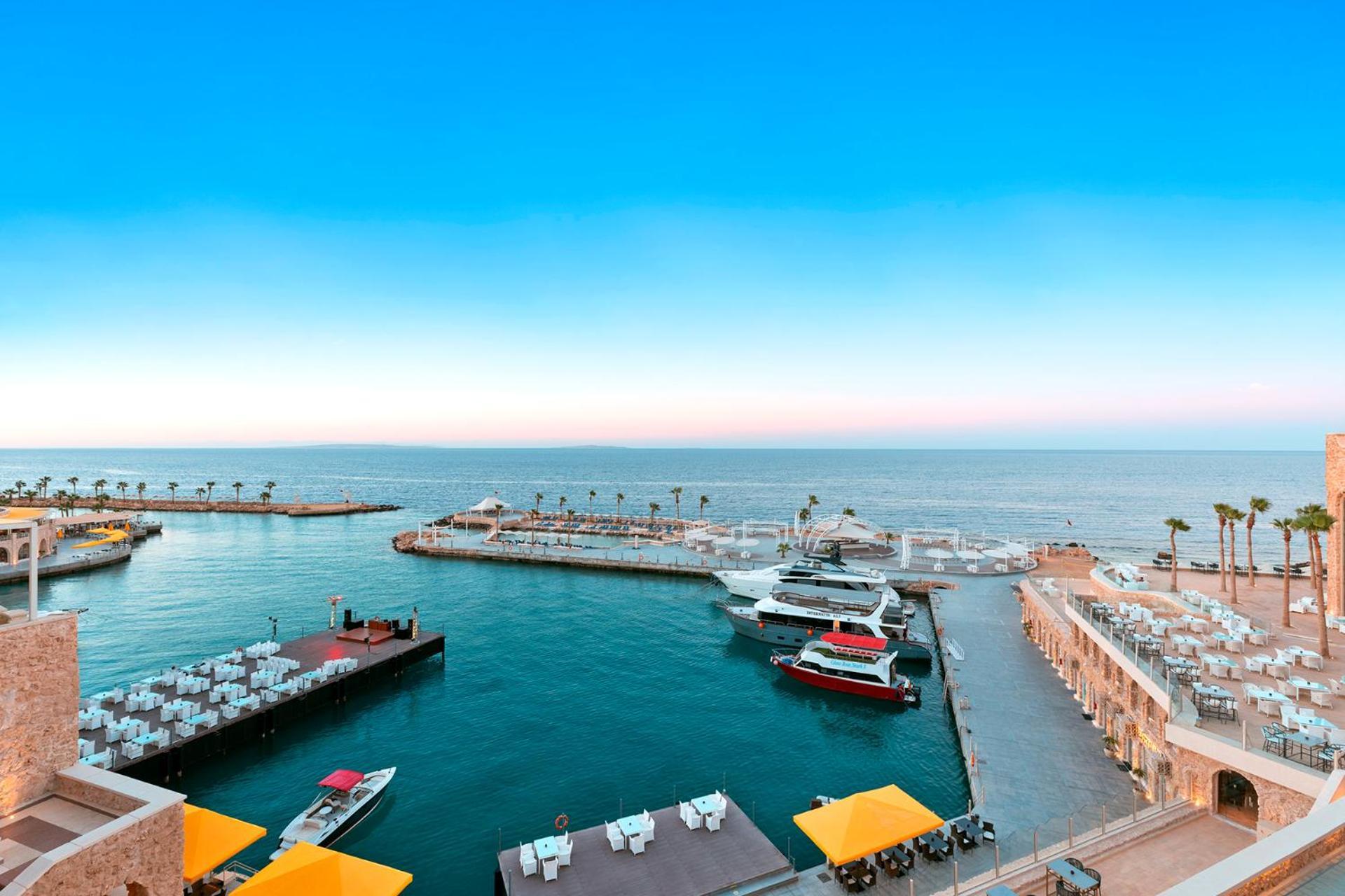 Pickalbatros Citadel Resort Sahl Hasheesh (Adults Only) Hurghada Exterior foto The photo shows a serene coastal scene featuring a marina. In the foreground, there are boats docked at a harbor, with some covered seating areas visible. The water is calm, reflecting the clear blue sky above, which transitions to a lighter hue clos