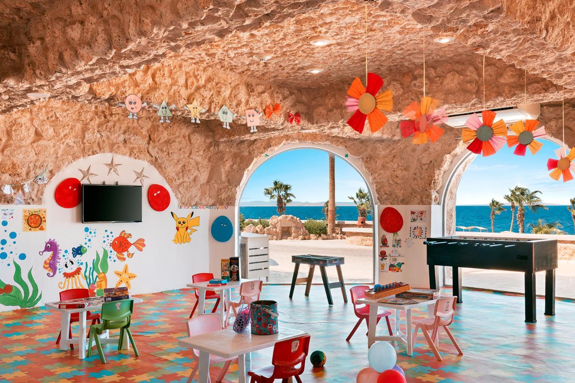 Pickalbatros Citadel Resort Sahl Hasheesh (Adults Only) Hurghada Exterior foto The photo depicts a vibrant children's playroom with a playful and colorful design. The room features a stone wall and large arched windows that provide a view of the ocean. Inside, there are several small tables and chairs in various bright colors, 
