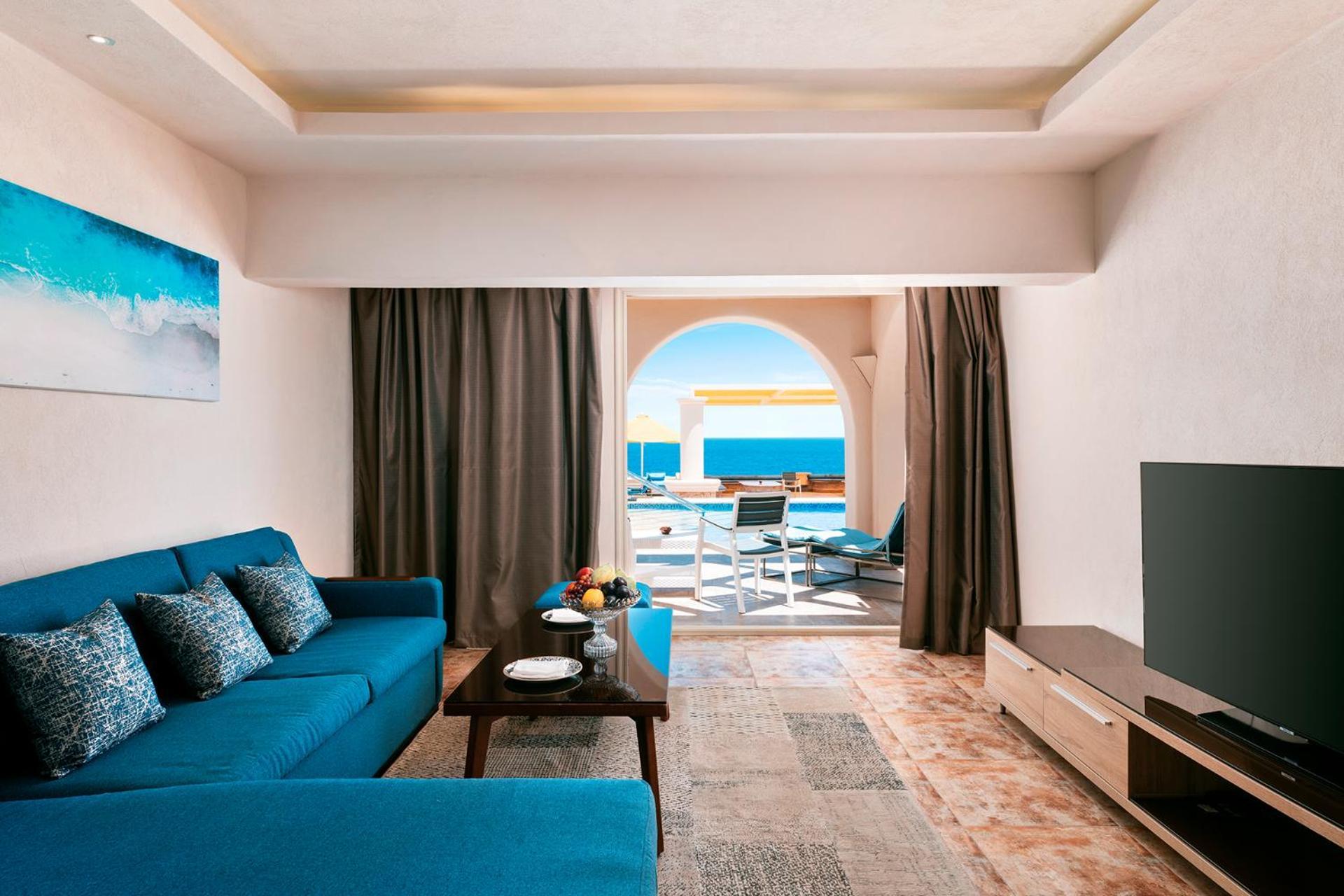 Pickalbatros Citadel Resort Sahl Hasheesh (Adults Only) Hurghada Exterior foto The photo shows a modern and stylish living room area. There is a teal sofa with several decorative cushions, a coffee table in front of it, and a television mounted on a light-colored entertainment unit. The room has large curtains that frame an ope