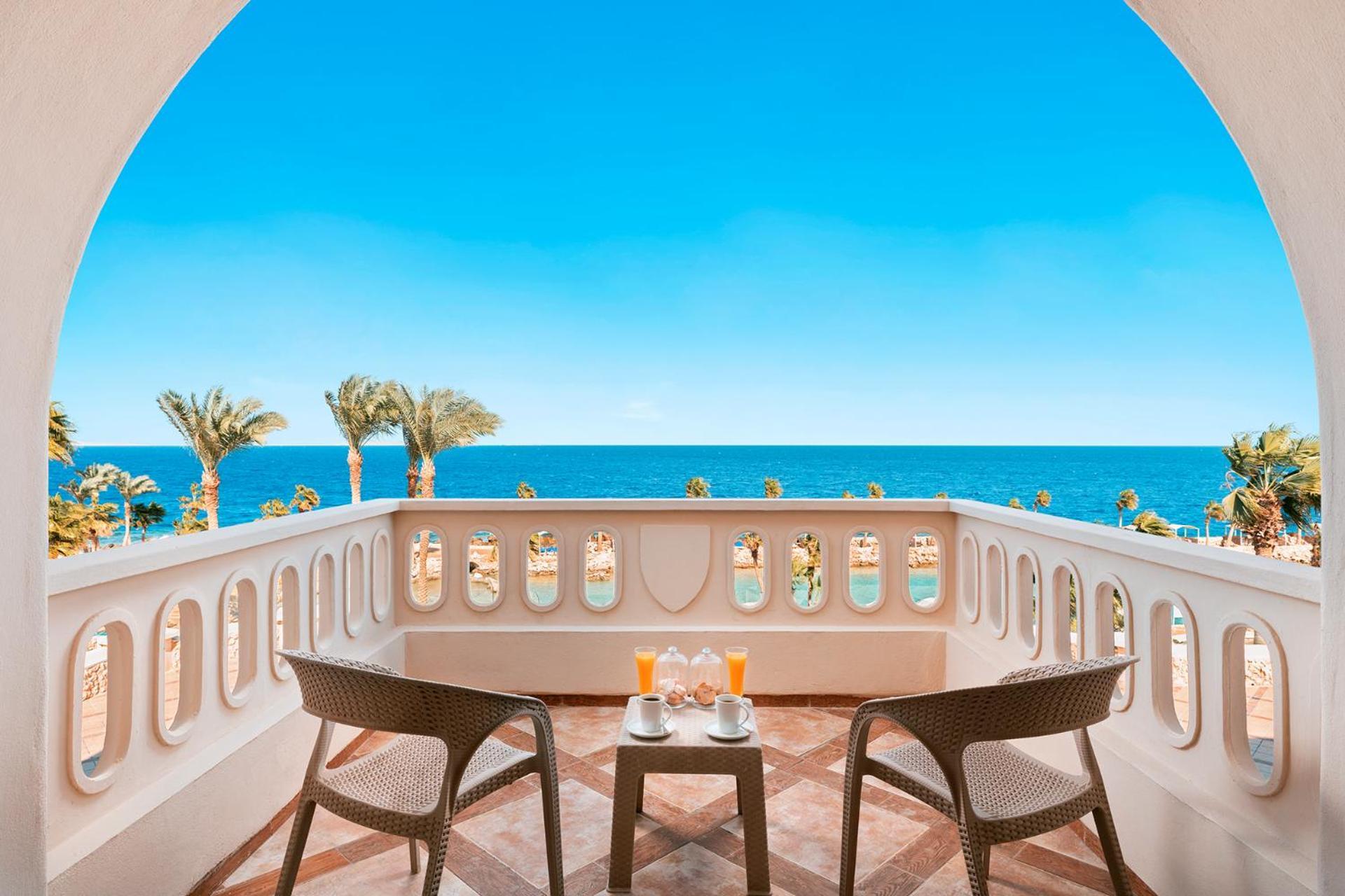 Pickalbatros Citadel Resort Sahl Hasheesh (Adults Only) Hurghada Exterior foto The photo shows a beautiful balcony overlooking the sea. The scene features two comfortable chairs placed at a small table, which has two glasses sitting on it. The background includes a clear blue sky and the ocean, with palm trees visible to the si