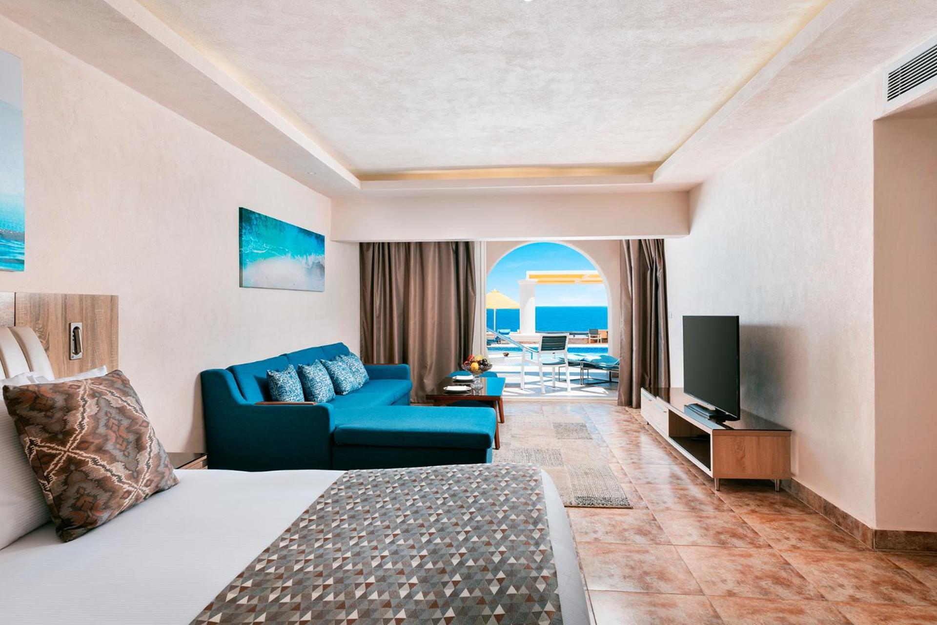 Pickalbatros Citadel Resort Sahl Hasheesh (Adults Only) Hurghada Exterior foto The photo shows a spacious hotel room featuring a comfortable bed with a patterned blanket. On one side, there is a stylish blue sofa with decorative pillows and a coffee table nearby. A flat-screen TV is mounted on a media unit facing the bed. Large
