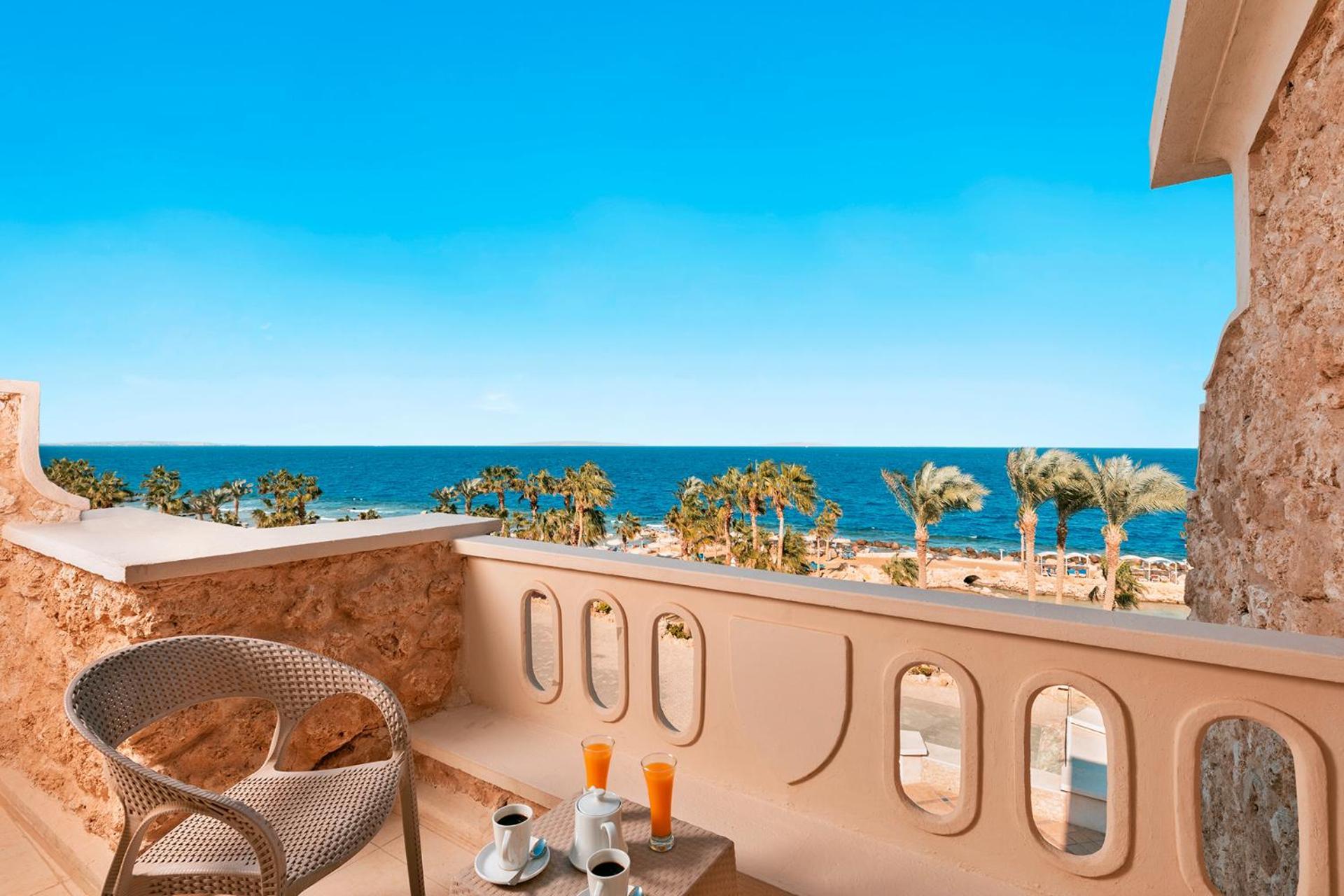 Pickalbatros Citadel Resort Sahl Hasheesh (Adults Only) Hurghada Exterior foto The photo depicts a balcony overlooking a beautiful ocean view. In the foreground, there is a small table with two cups, possibly of coffee, and two glasses of what appears to be orange juice. A comfortable woven chair sits beside the table. The back