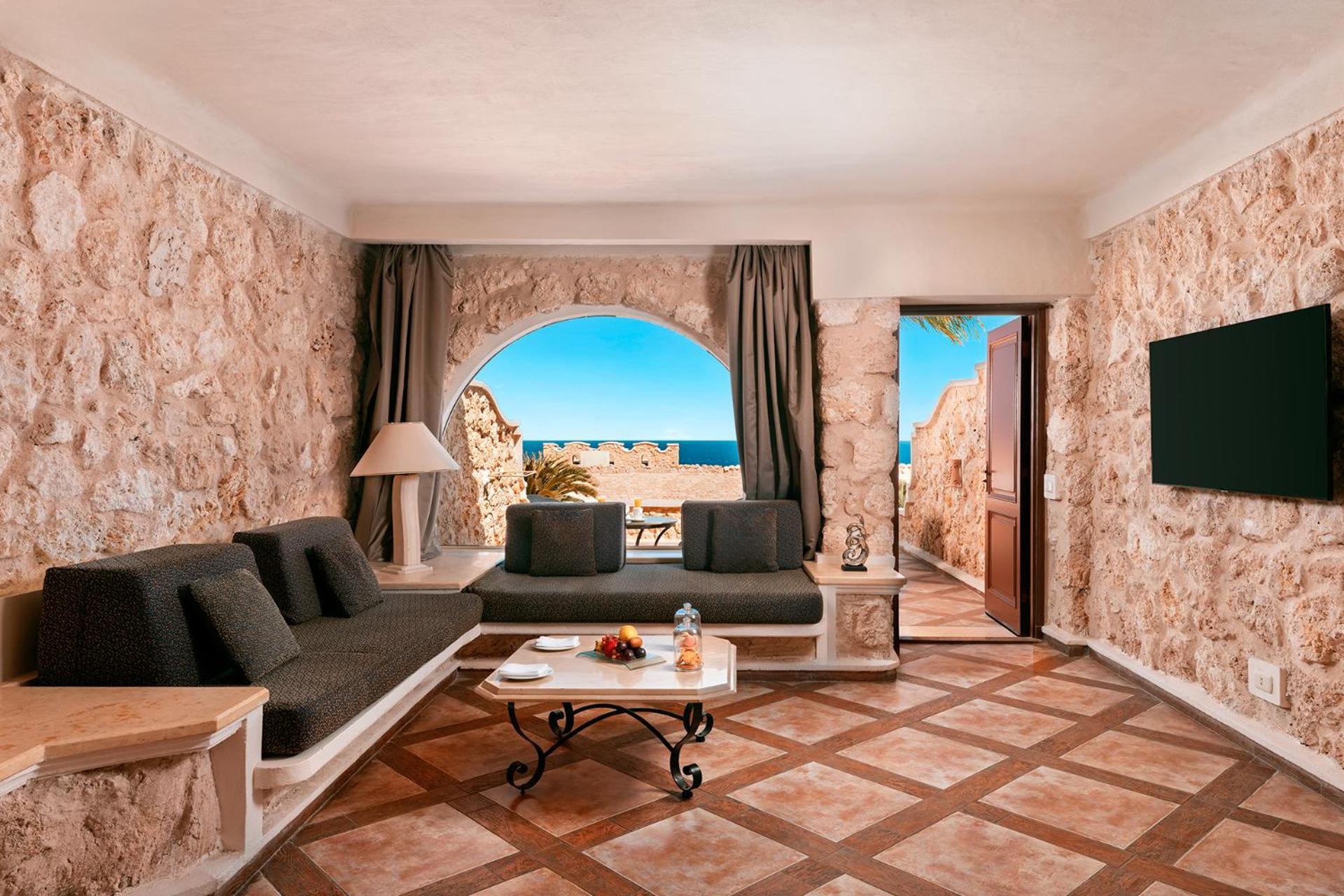 Pickalbatros Citadel Resort Sahl Hasheesh (Adults Only) Hurghada Exterior foto The photo shows an inviting living space with a coastal view. The room features stone walls, giving it a rustic charm. There's a comfortable seating area with dark gray sofas arranged around a stylish coffee table. Natural light floods the room throu