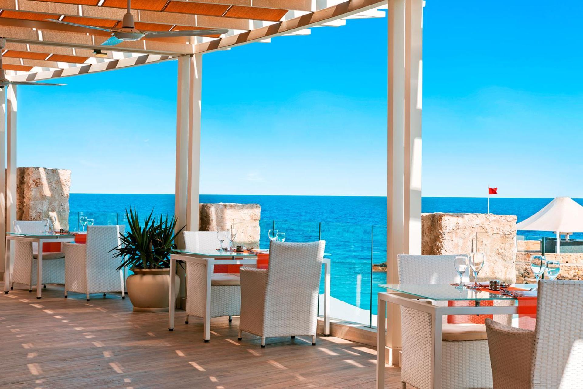 Pickalbatros Citadel Resort Sahl Hasheesh (Adults Only) Hurghada Exterior foto The photo shows a modern restaurant with a covered terrace overlooking a bright blue sea. The space features white tables and chairs, with some greenery in decorative pots. The scene captures a clear sky with minimal clouds, suggesting a sunny day. O
