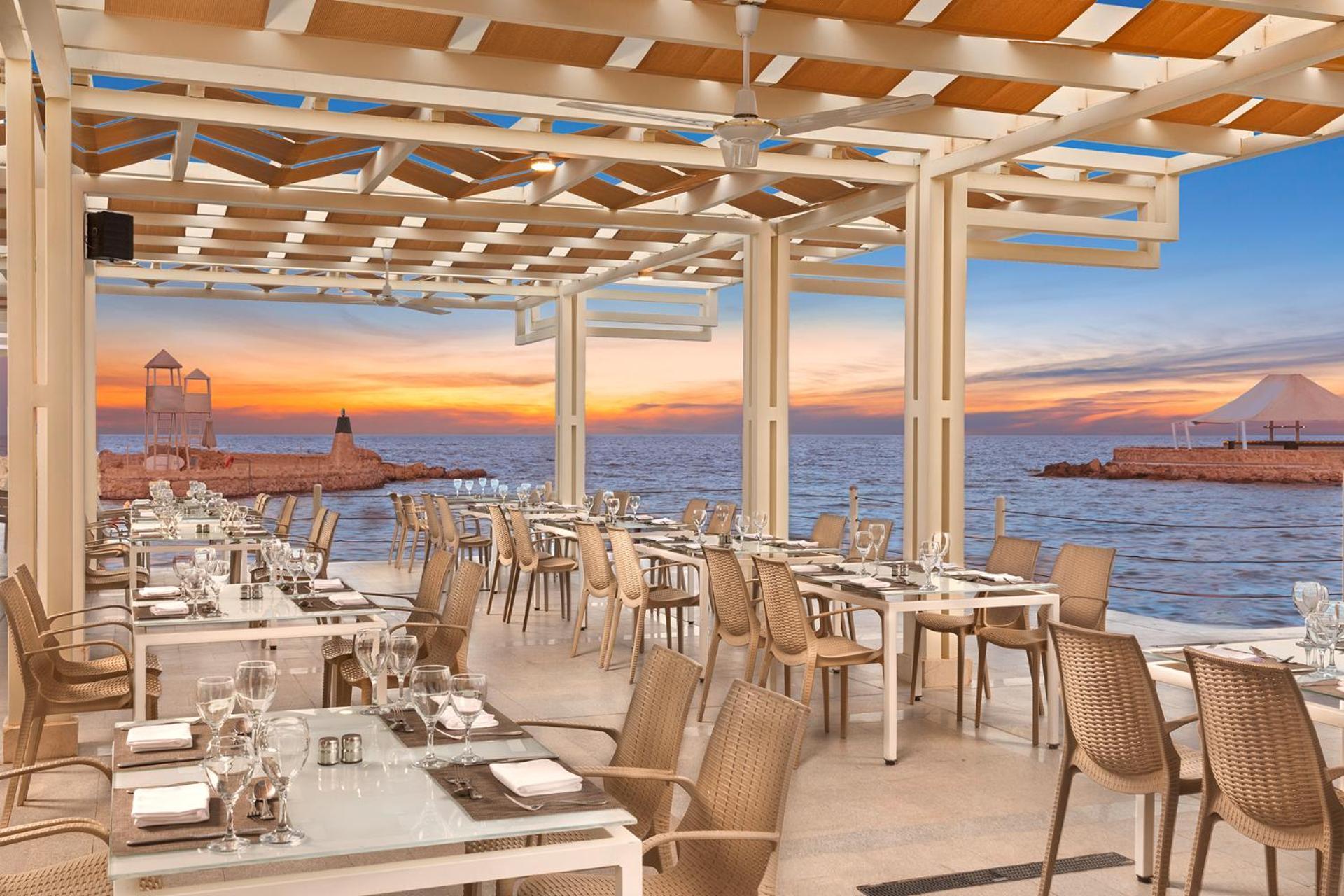Pickalbatros Citadel Resort Sahl Hasheesh (Adults Only) Hurghada Exterior foto The photo shows a modern restaurant with an outdoor seating area. The design features a pergola with a wooden roof that lets in natural light. There are several white tables set with cutlery and glassware, indicating readiness for dining. In the back