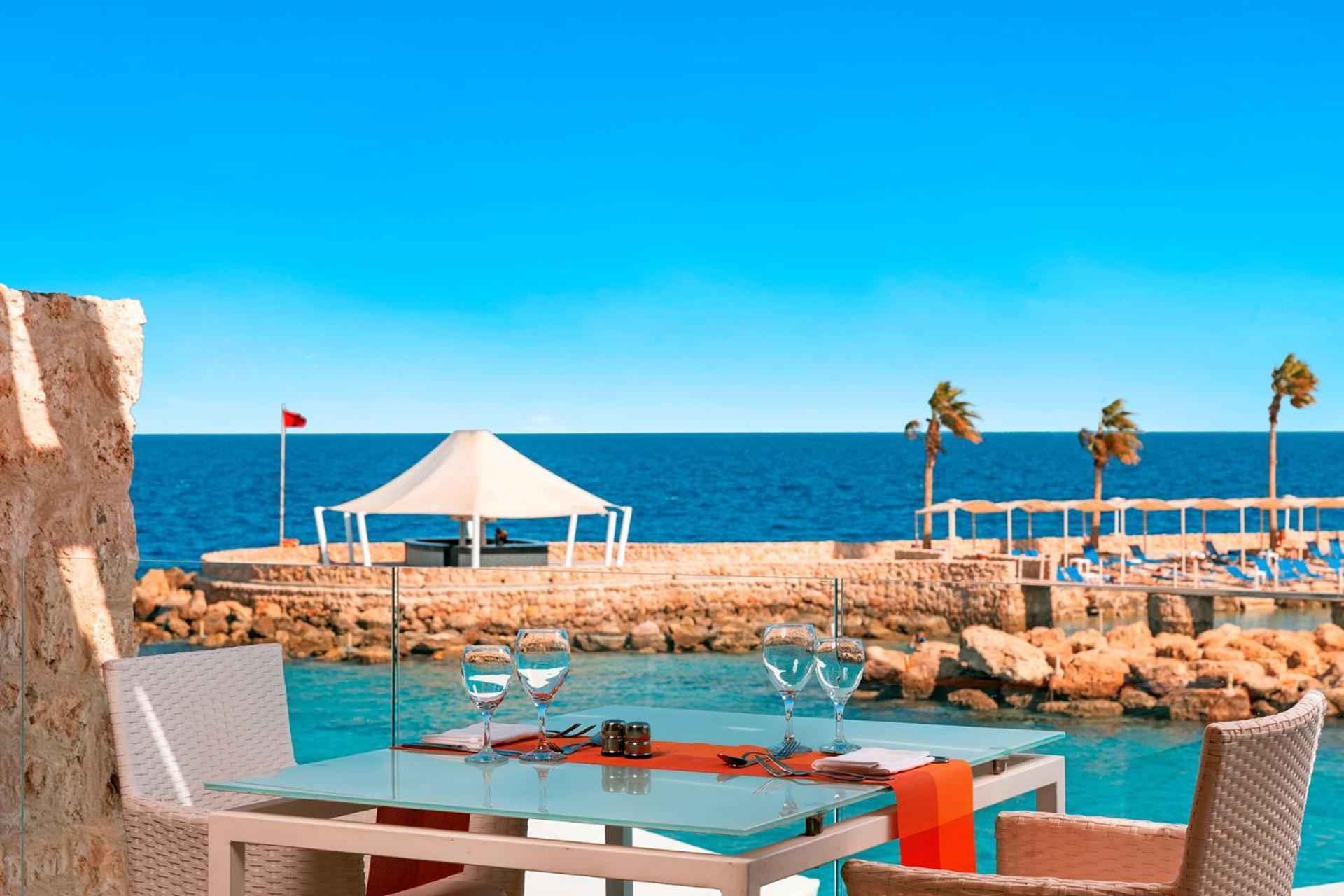 Pickalbatros Citadel Resort Sahl Hasheesh (Adults Only) Hurghada Exterior foto The photo depicts a serene seaside view from a restaurant table. There's a glass table set with two wine glasses and a central decorative item. In the background, you can see calm blue waters and a coastline with palm trees. A white canopy or tent is