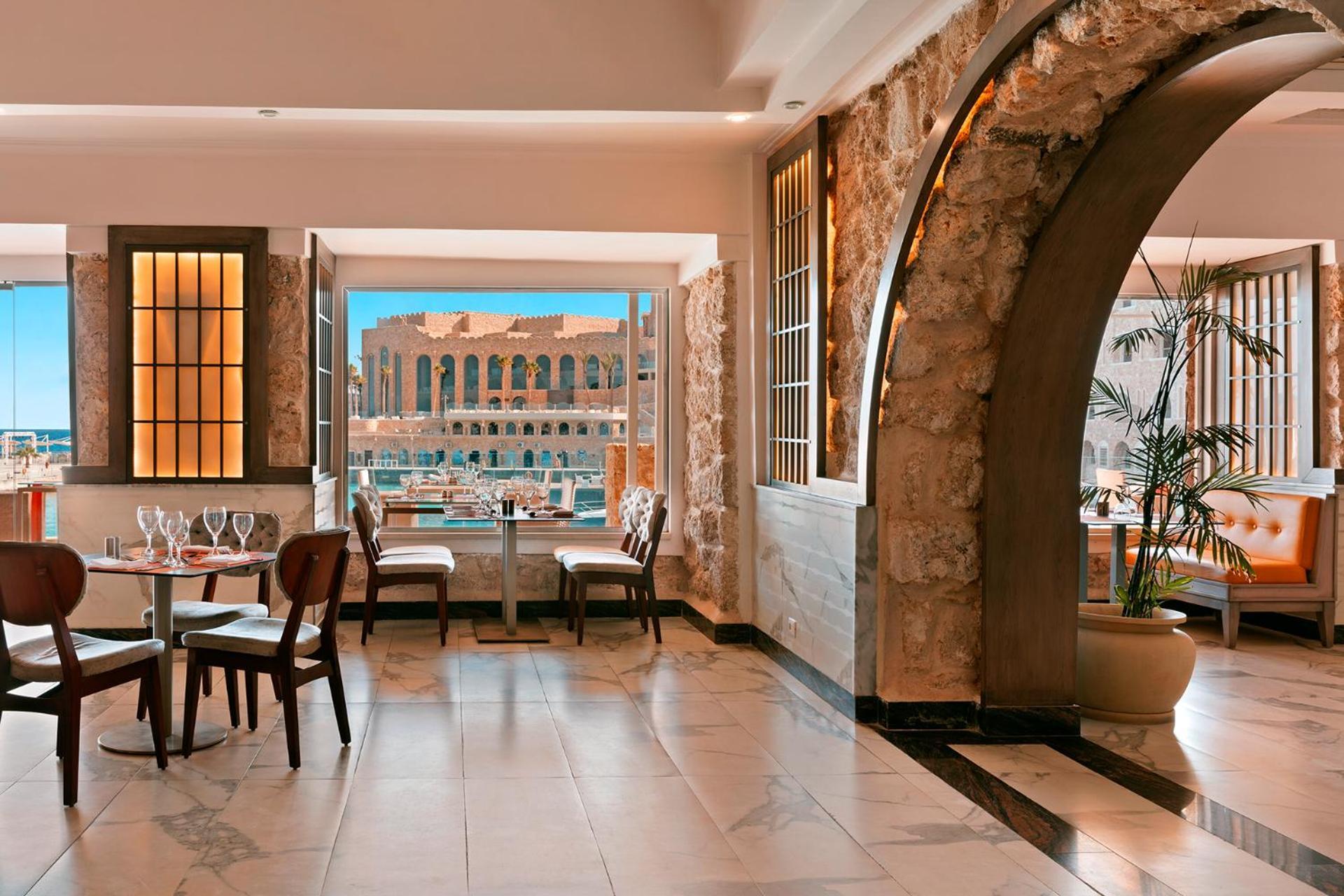 Pickalbatros Citadel Resort Sahl Hasheesh (Adults Only) Hurghada Exterior foto The photo depicts a beautifully designed restaurant interior. It features elegant wooden tables and chairs set up near large windows that open to a scenic view. The walls are a combination of stone and smooth finishes, creating a warm and inviting at
