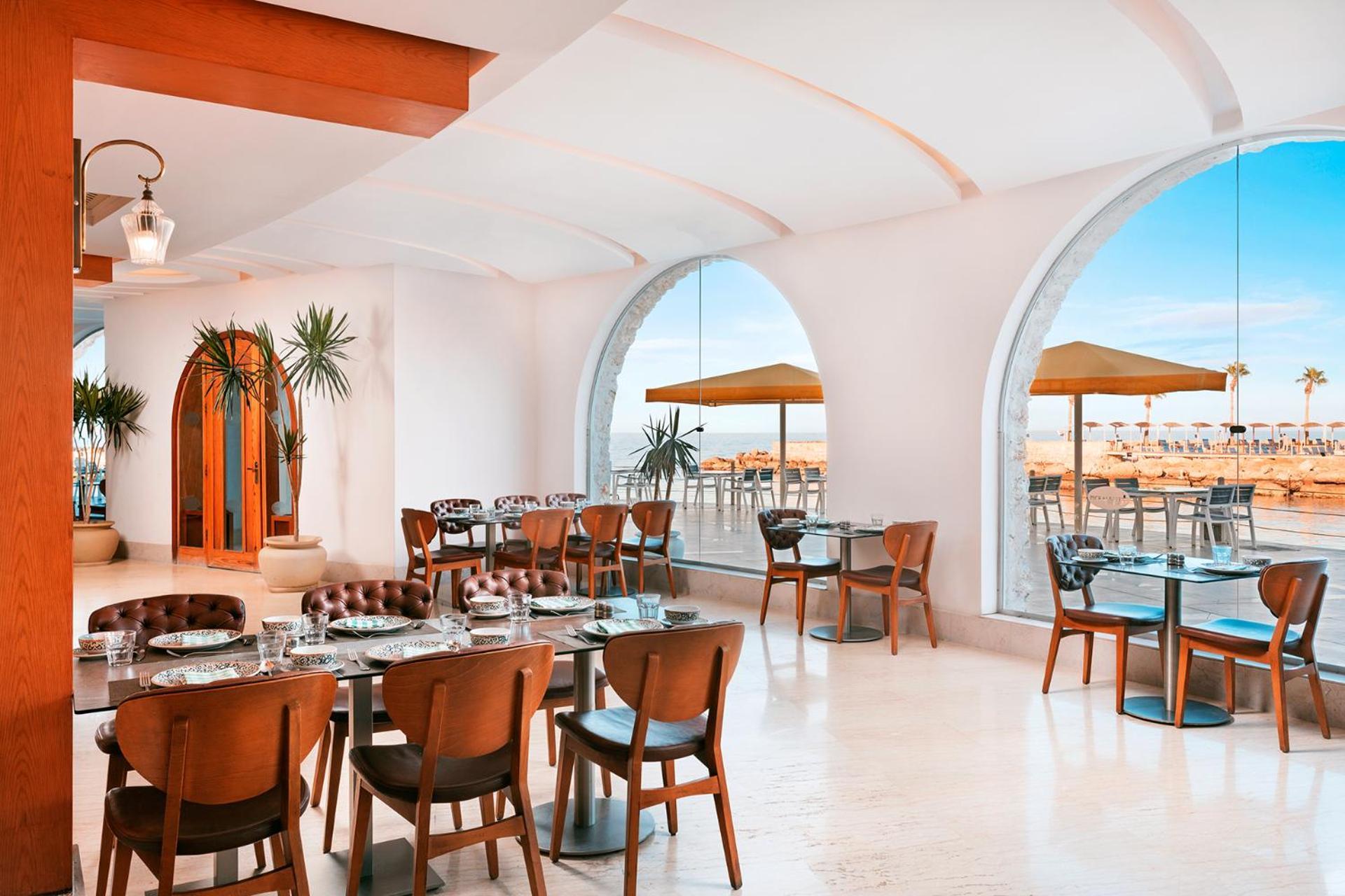 Pickalbatros Citadel Resort Sahl Hasheesh (Adults Only) Hurghada Exterior foto The photo shows a modern restaurant interior featuring elegant wooden furniture and a bright, airy atmosphere. There are several tables with chairs arranged in a spacious layout. Large arched windows allow natural light to flood the space, and throug