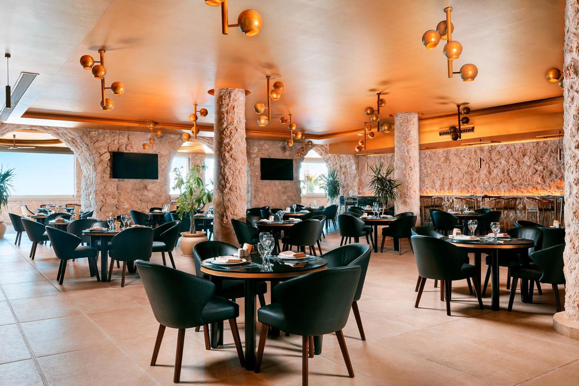 Pickalbatros Citadel Resort Sahl Hasheesh (Adults Only) Hurghada Exterior foto The photo shows a modern restaurant interior. It features elegant round tables with black chairs arranged in a spacious layout. The walls are adorned with a textured, stone-like finish, and there are large windows that likely offer a view outside. Th