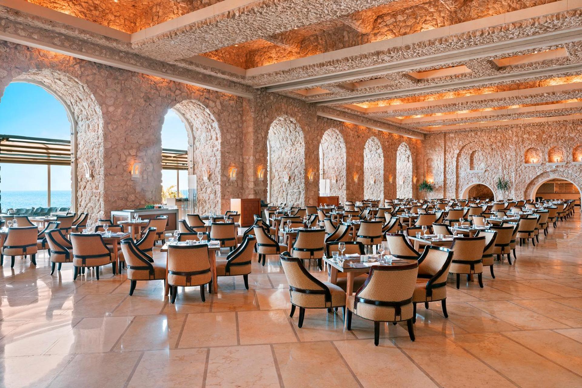 Pickalbatros Citadel Resort Sahl Hasheesh (Adults Only) Hurghada Exterior foto The photo shows an elegant restaurant interior featuring a spacious layout with numerous dining tables and chairs. The room is adorned with beautiful stone walls and large arched windows that allow natural light to flood in, offering a view of the ex