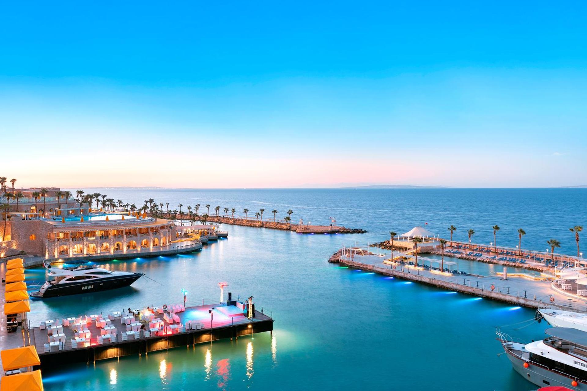 Pickalbatros Citadel Resort Sahl Hasheesh (Adults Only) Hurghada Exterior foto The photo depicts a tranquil coastal scene featuring a marina and a beautiful seascape. There are several yachts and boats anchored in the calm, turquoise waters. Along the shore, there are palm trees and a well-lit area that seems to host dining or 