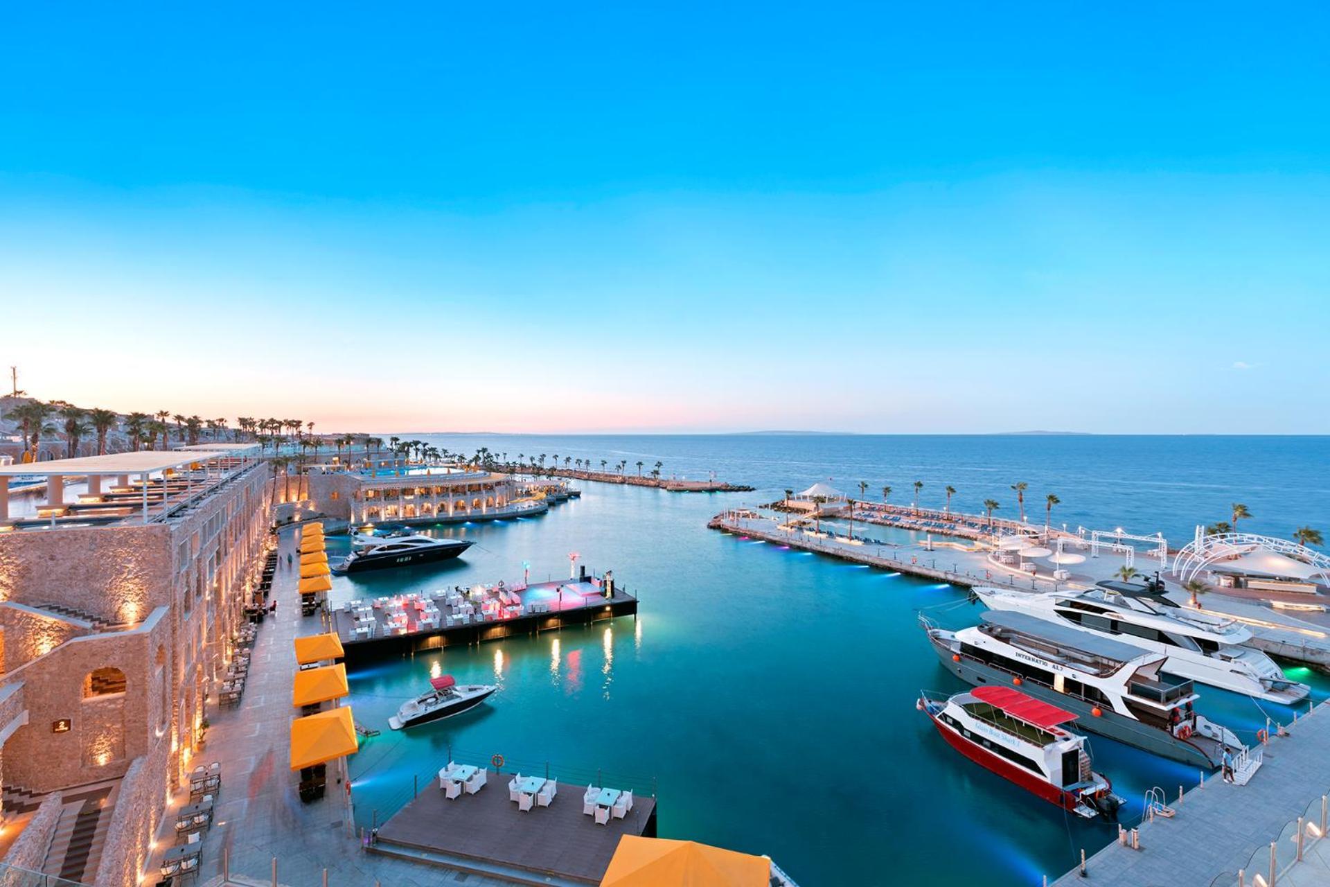 Pickalbatros Citadel Resort Sahl Hasheesh (Adults Only) Hurghada Exterior foto The photo depicts a picturesque harbor scene at sunset. The marina features several boats and yachts docked along the water, with some colorful boats visible. There are also areas with lounge chairs and umbrellas, suggesting a relaxing atmosphere. Th