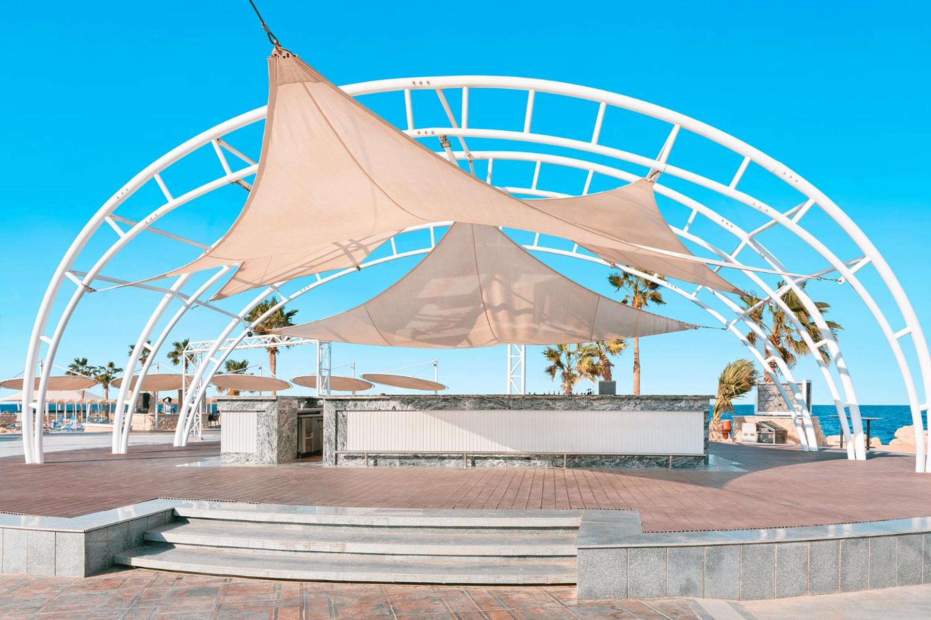 Pickalbatros Citadel Resort Sahl Hasheesh (Adults Only) Hurghada Exterior foto The photo shows an open-air structure with a modern design, featuring large sail-like fabric canopies suspended above a raised stage or platform. The structure is supported by a white metal framework and is set against a bright blue sky. In the backg
