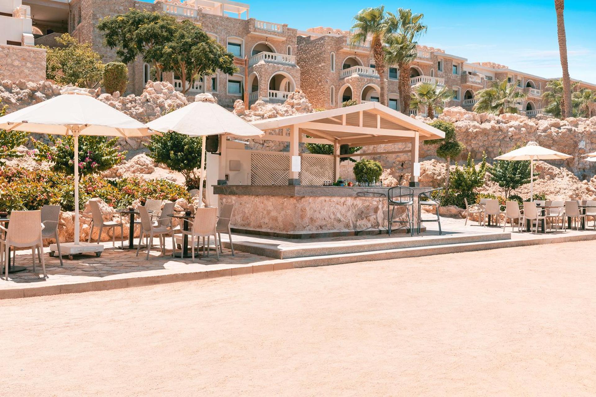 Pickalbatros Citadel Resort Sahl Hasheesh (Adults Only) Hurghada Exterior foto The photo depicts a beautiful outdoor area, likely at a resort. In the foreground, there is a bar or food service area with a countertop. Surrounding the bar, there are several tables with chairs, some of which are shaded by large umbrellas. In the b
