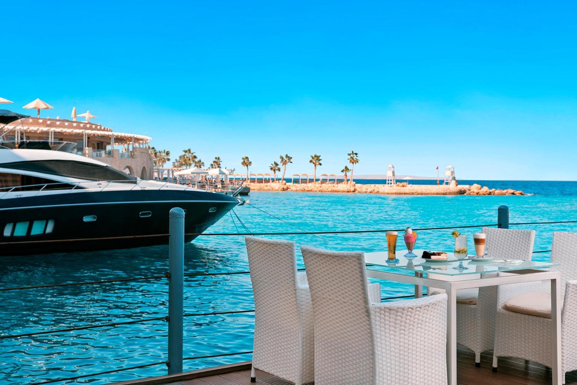 Pickalbatros Citadel Resort Sahl Hasheesh (Adults Only) Hurghada Exterior foto The photo shows a beautiful waterfront scene. In the foreground, there is a set of four white wicker chairs around a glass table, which is set with colorful drinks. In the background, you can see a luxurious yacht docked at the edge of a clear blue s