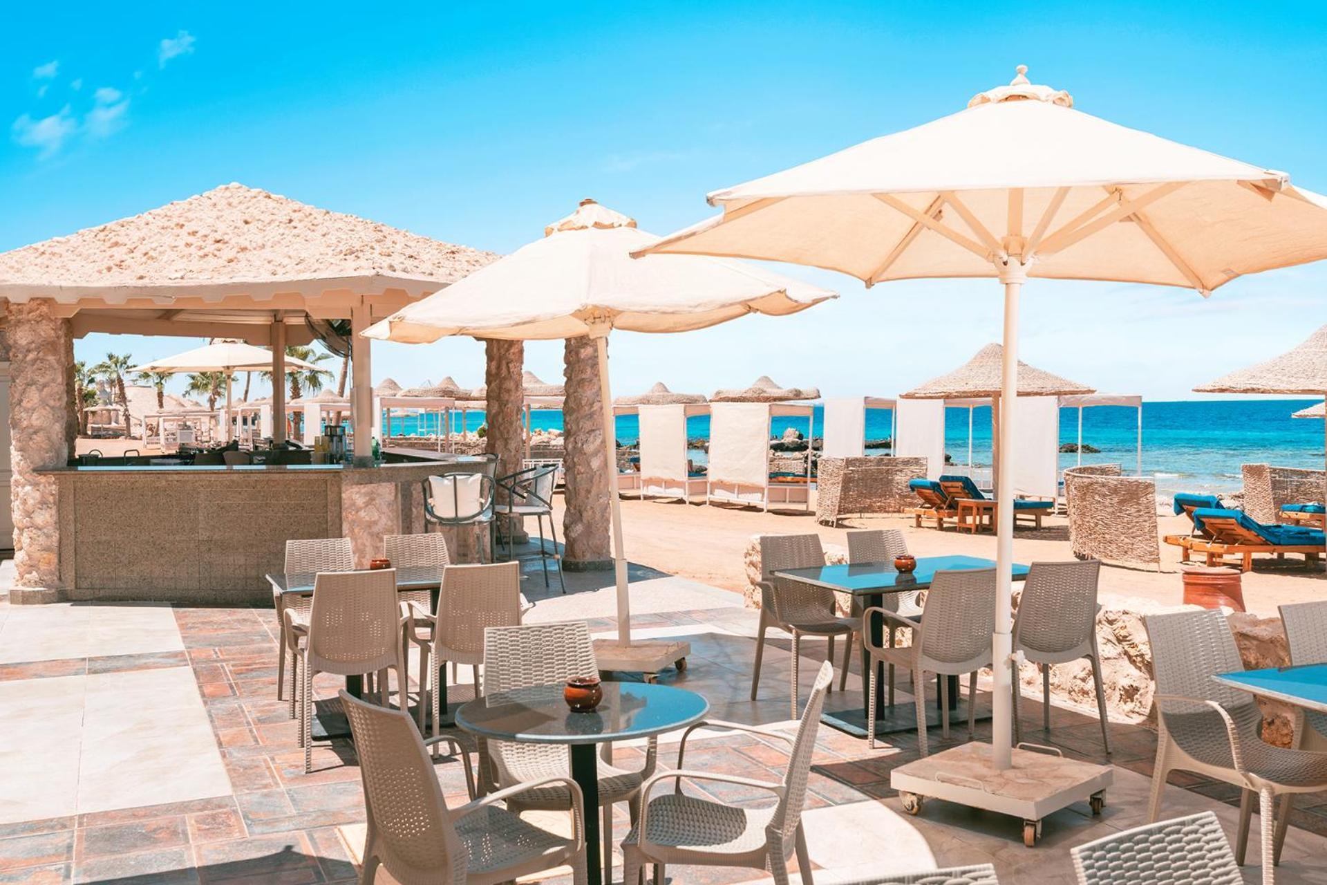 Pickalbatros Citadel Resort Sahl Hasheesh (Adults Only) Hurghada Exterior foto The photo depicts a beachside area with several tables and chairs arranged neatly. There are large umbrellas providing shade over the tables, creating a cozy atmosphere for dining or relaxing. In the background, there are sun loungers and umbrellas s