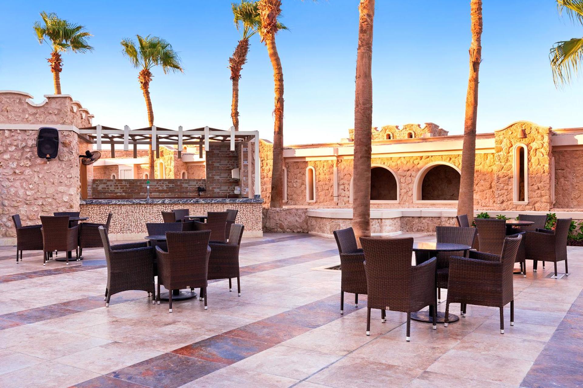 Pickalbatros Citadel Resort Sahl Hasheesh (Adults Only) Hurghada Exterior foto The photo shows an outdoor seating area, likely part of a resort or restaurant. There are several tables and chairs arranged on a tiled patio, with palm trees in the background. The architecture includes stone walls and arches, giving a warm, invitin