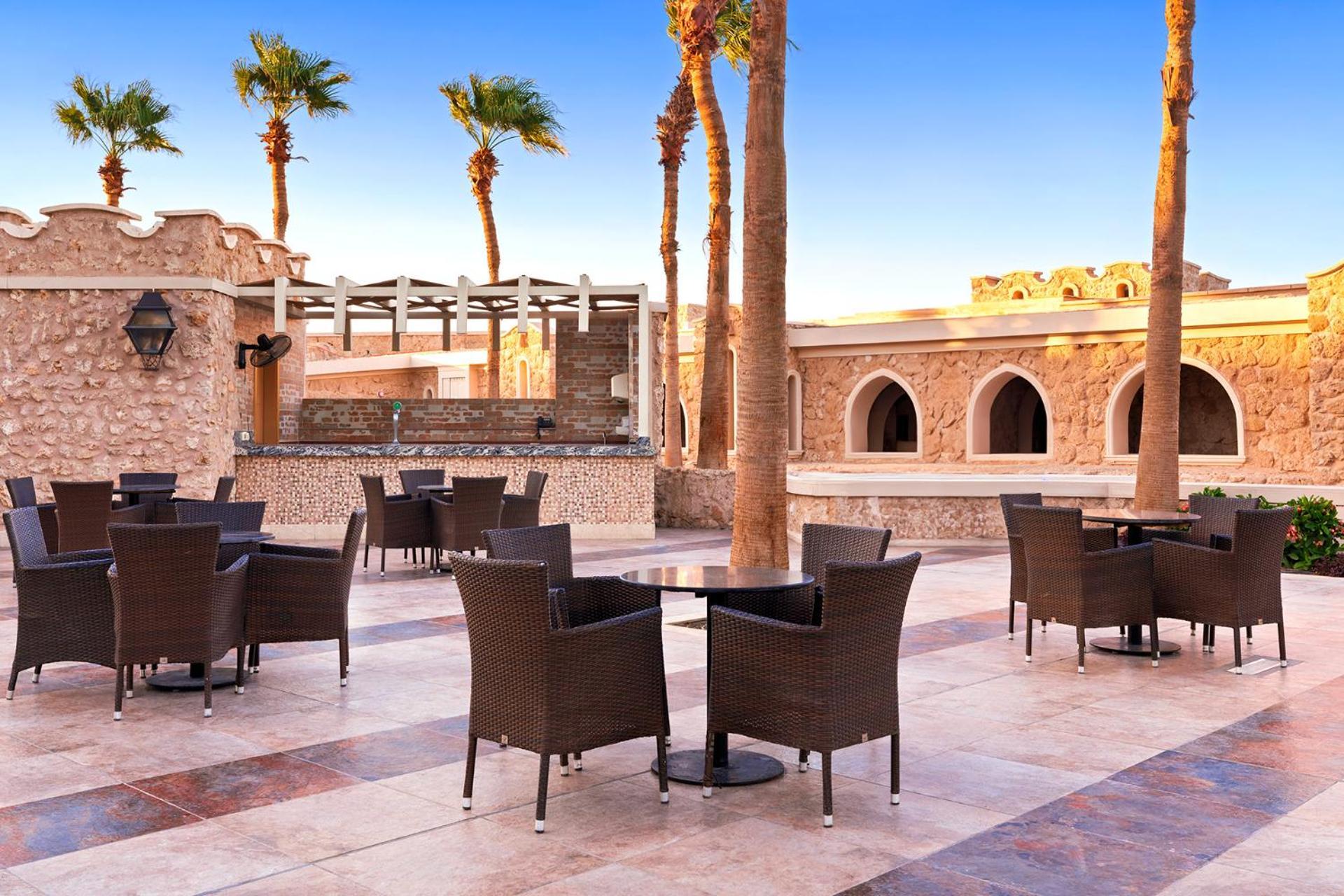 Pickalbatros Citadel Resort Sahl Hasheesh (Adults Only) Hurghada Exterior foto The photo shows an outdoor seating area with several tables and chairs arranged on a tiled patio. The furniture is made of dark wicker, and each table has a single chair positioned around it. Palm trees rise prominently in the background, adding a tr