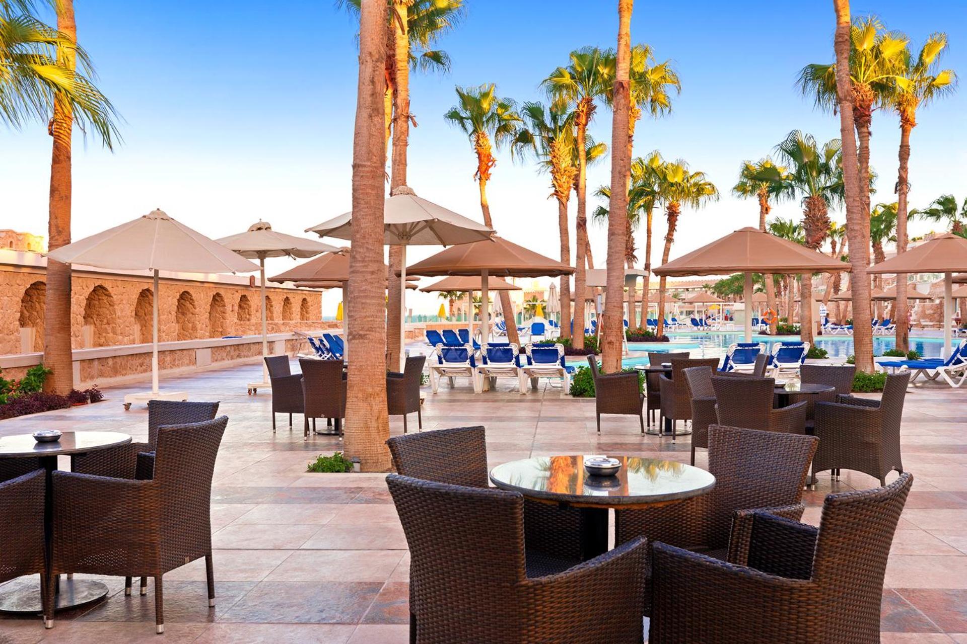 Pickalbatros Citadel Resort Sahl Hasheesh (Adults Only) Hurghada Exterior foto The photo depicts a sunny outdoor scene at a resort. There are several palm trees lining the area, providing a tropical atmosphere. In the foreground, there are stylish wicker chairs and tables set up, possibly for dining or relaxation. In the backgr