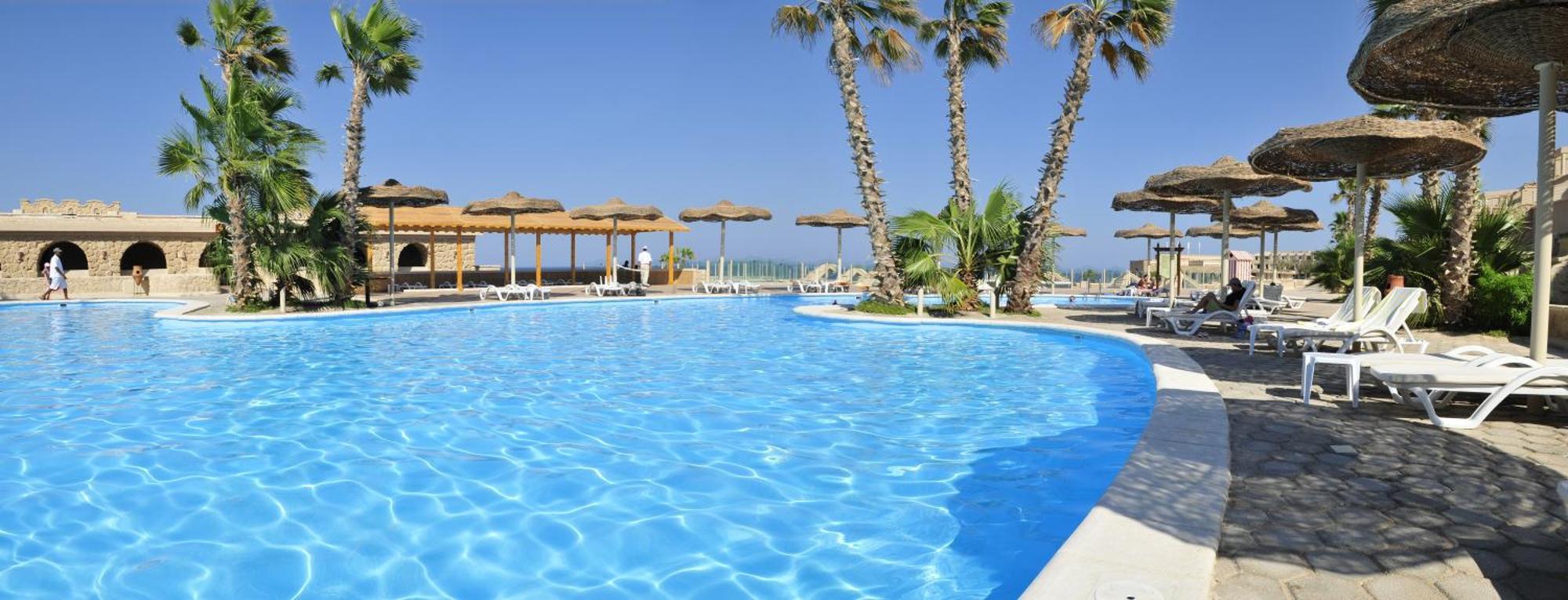 Pickalbatros Citadel Resort Sahl Hasheesh (Adults Only) Hurghada Exterior foto The photo shows a picturesque pool area surrounded by palm trees and lounge chairs. The swimming pool has a clear blue water, creating a refreshing atmosphere. In the background, there are thatched umbrellas providing shade, likely indicating a tropi