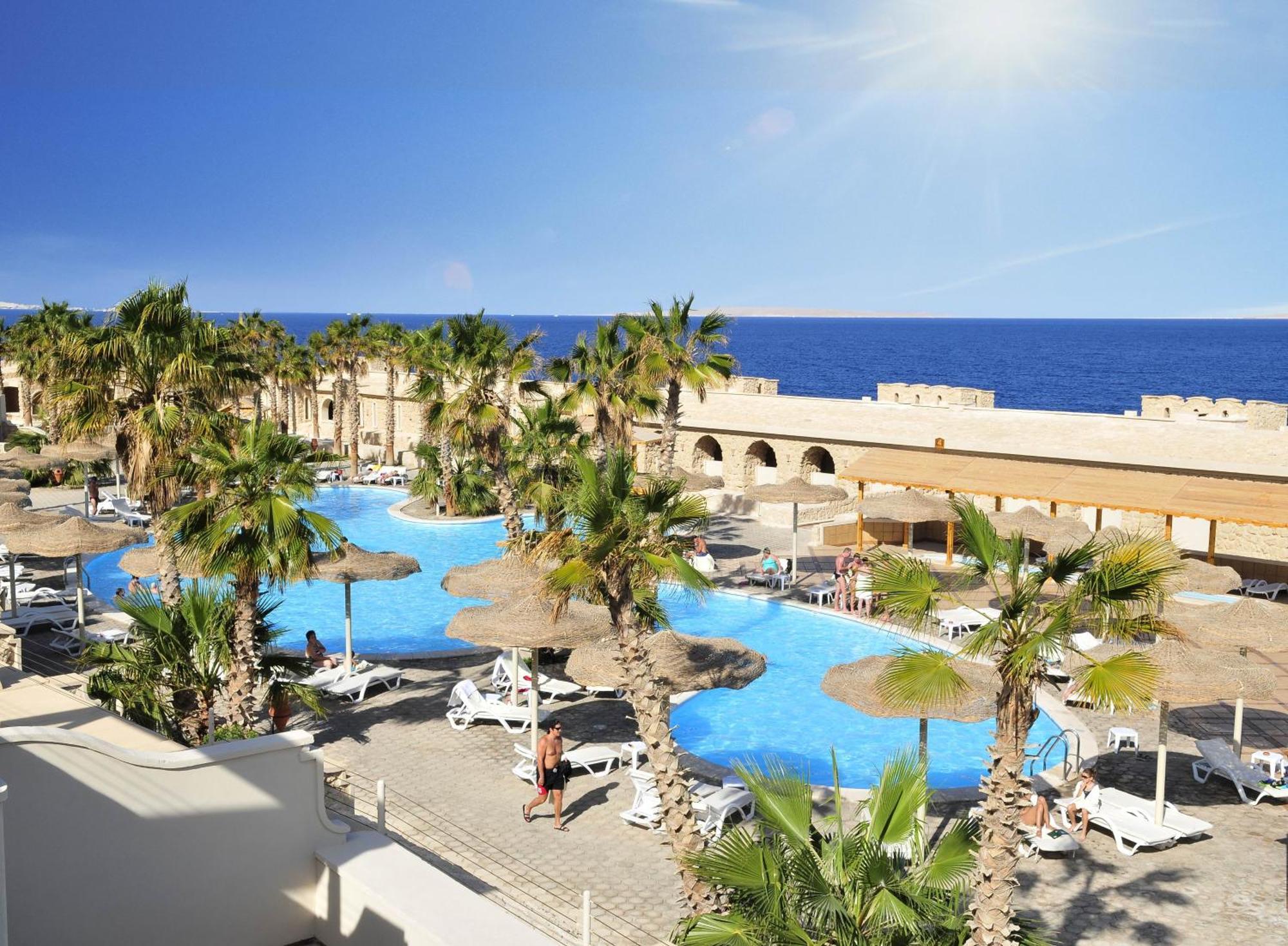 Pickalbatros Citadel Resort Sahl Hasheesh (Adults Only) Hurghada Exterior foto The photo shows a sunny resort area featuring a large swimming pool surrounded by lush palm trees. The pool is blue and has several lounge chairs and umbrellas set up along its edges, suggesting a relaxing atmosphere. In the background, there is a vi